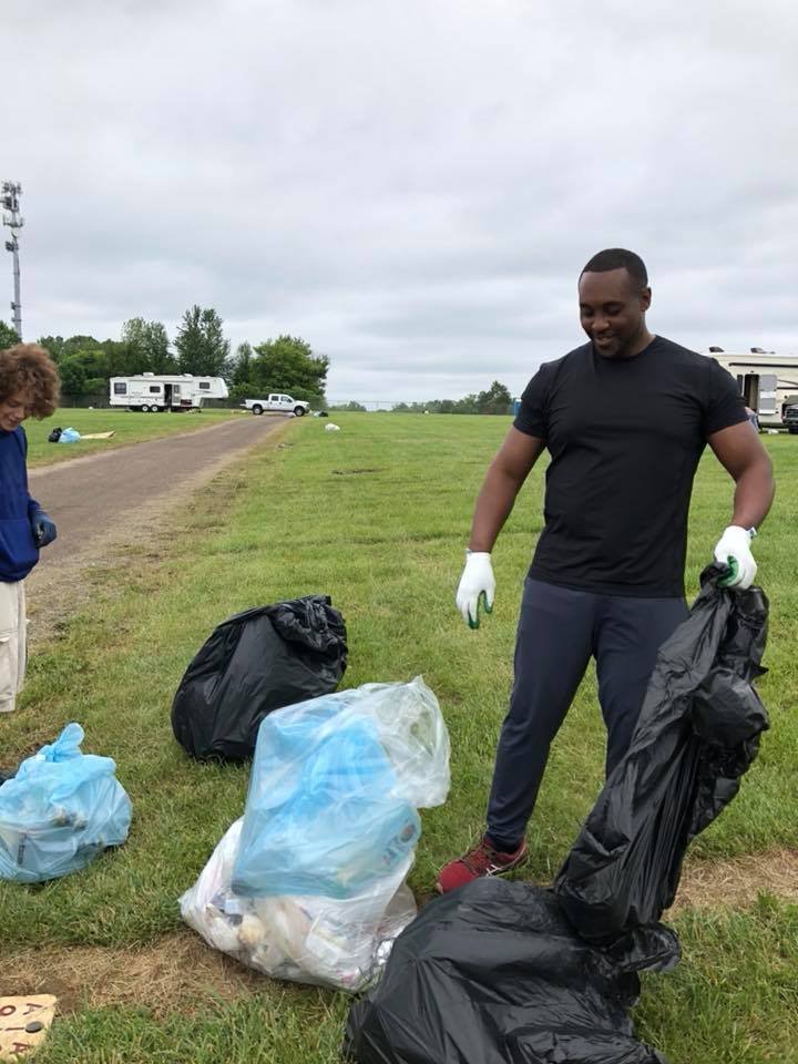 Two people are picking up trash in a grassy area near a dirt path. The man in the foreground is holding a large black trash bag and wearing gloves. The woman is standing nearby with another trash bag. In the background, there are several caravans and