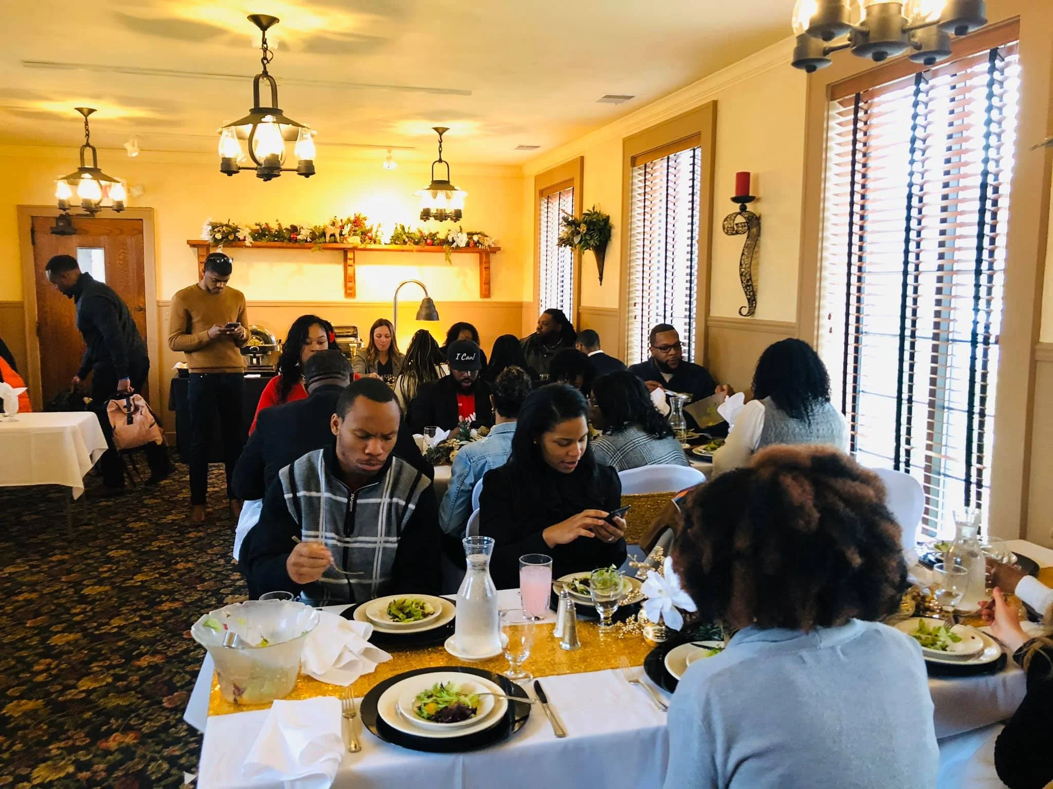 A group of people sitting at a dining table in a restaurant or banquet hall, with plate of salads and glasses of water. Several people are engaged with their phones or talking, and two servers are attending to the guests.