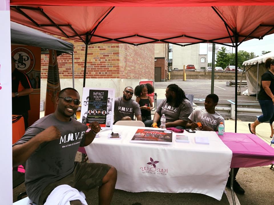 Group of people sitting at a table under a pink canopy tent at an outdoor event. They are wearing matching gray T-shirts with 