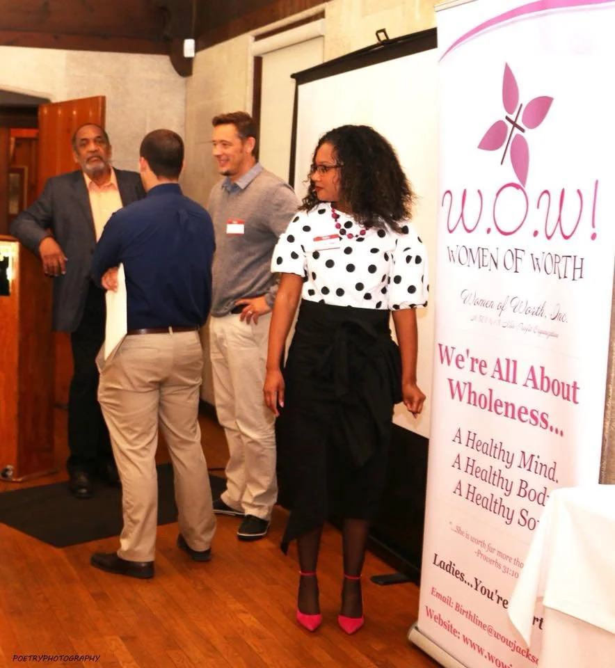 Four people are standing in front of a white screen and a banner that says "W.O.W! Women of Worth" at a wellness event. The group includes three men and one woman, who is dressed in a polka dot blouse and black skirt. They are inside a wood-paneled r