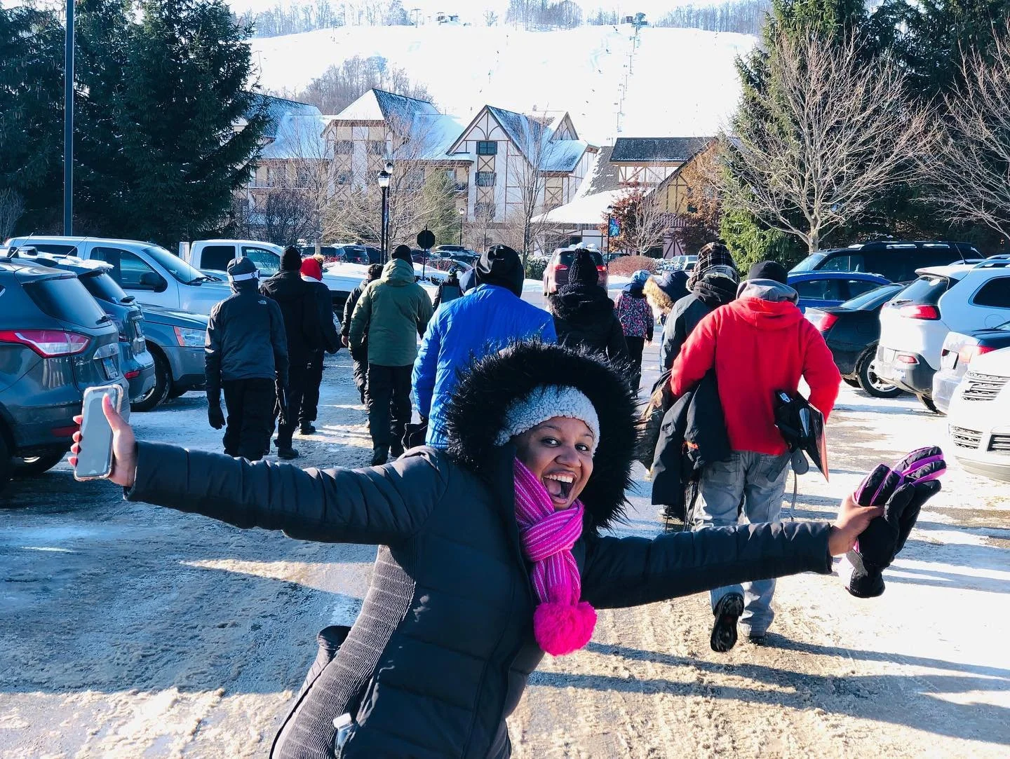A woman smiling and posing with her arms outstretched in a snowy parking lot with a group of people walking away in the background and snow-covered buildings and trees.