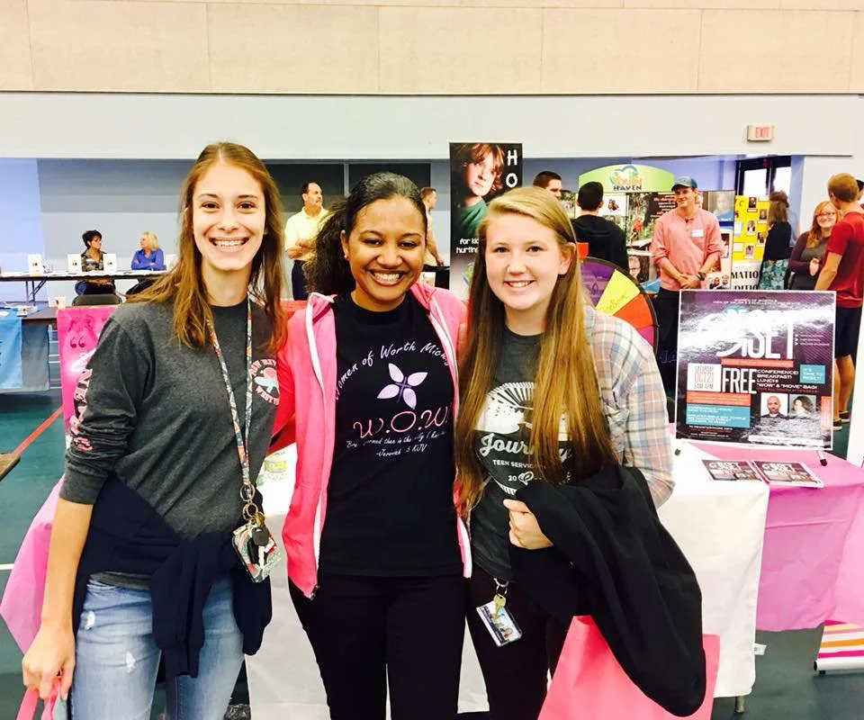 Three young women standing together at a conference booth, smiling, with various people and displays in the background.