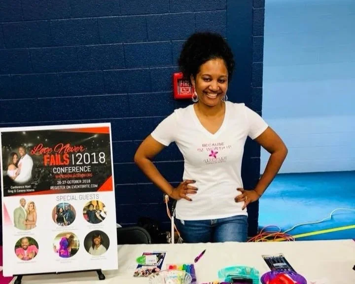 A woman standing behind a table at a conference, smiling and posing with hands on hips. She is wearing a white T-shirt and jeans, with a dark blue wall and a fire alarm in the background. The table has colorful items and a promotional sign for the Lo
