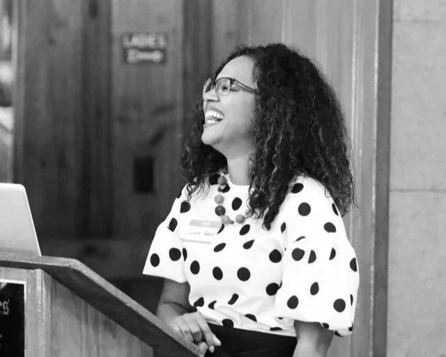 A woman with curly hair and glasses sitting at a desk, smiling and wearing a polka dot top, in a wood-paneled room.