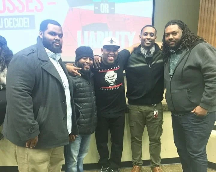 Group of five men standing arm-in-arm indoors, smiling at the camera, with a projector screen behind them showing a presentation.