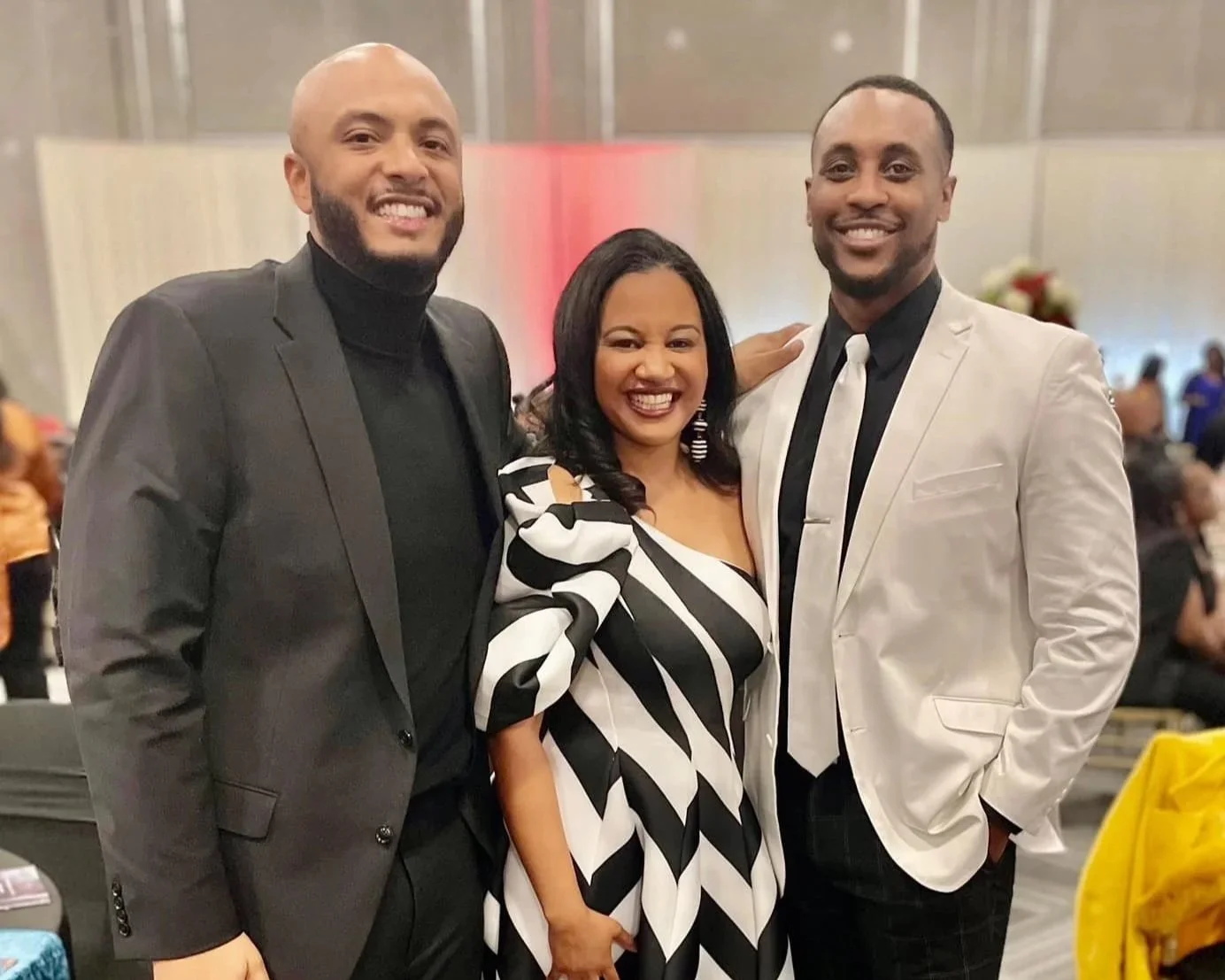 Three people smiling and posing for a photo at an indoor event, with a woman in the center wearing a black and white dress, flanked by two men in suits.