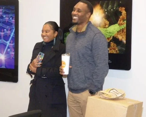 A woman and a man standing next to each other, smiling. The woman is holding a smartphone, and the man is holding a drink in a clear cup. They are indoors, with two digital screens displaying colorful images behind them.