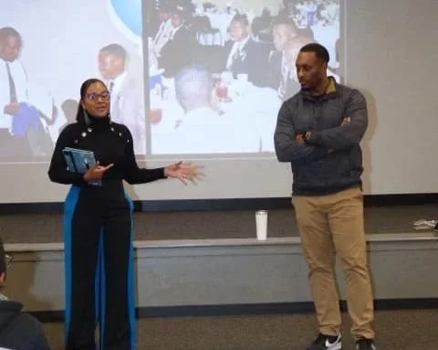 A woman is speaking or presenting while a man listens in a room with a large projected screen showing a group of people in the background.