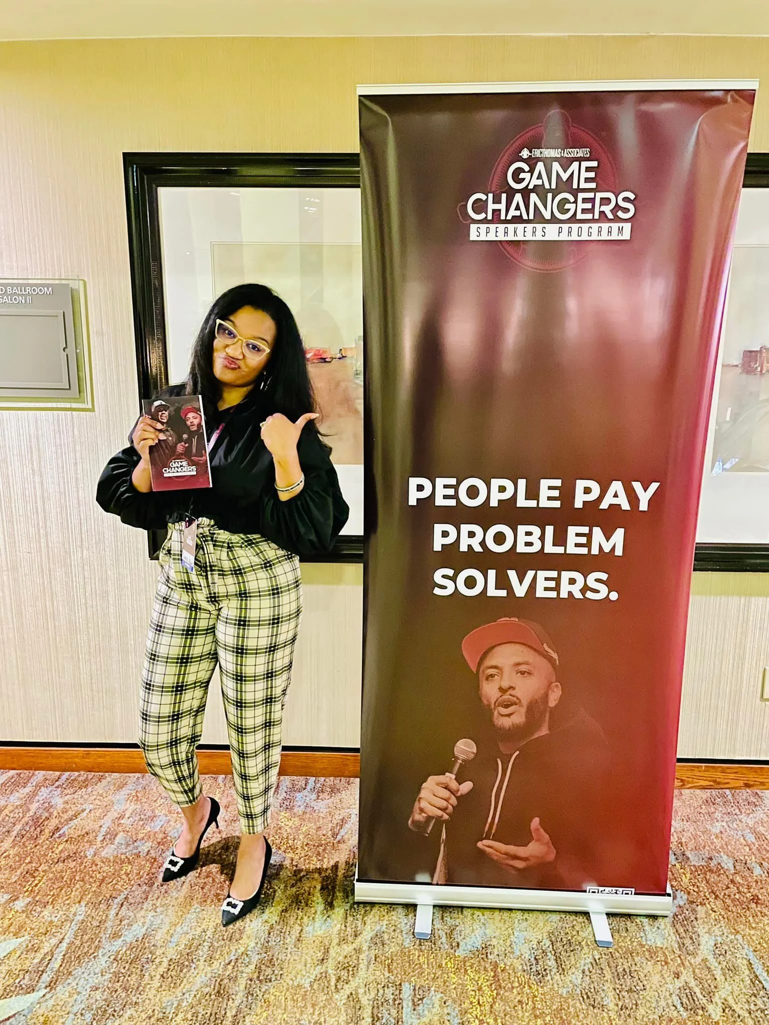 A woman standing next to a banner at an event, holding a booklet, dressed in a black top, plaid pants, and heels, pointing at the banner which reads 'People pay problem solvers' and features a man holding a microphone.