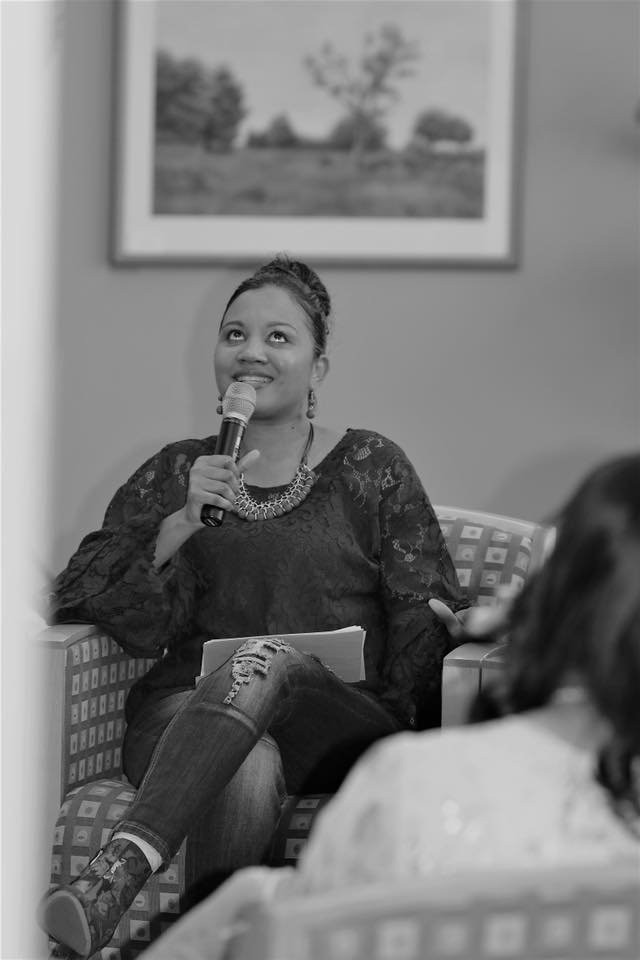 A woman sitting in a chair, holding a microphone and reviewing papers, looking up thoughtfully during a discussion or interview, with a framed picture on the wall behind her and other people in the foreground.