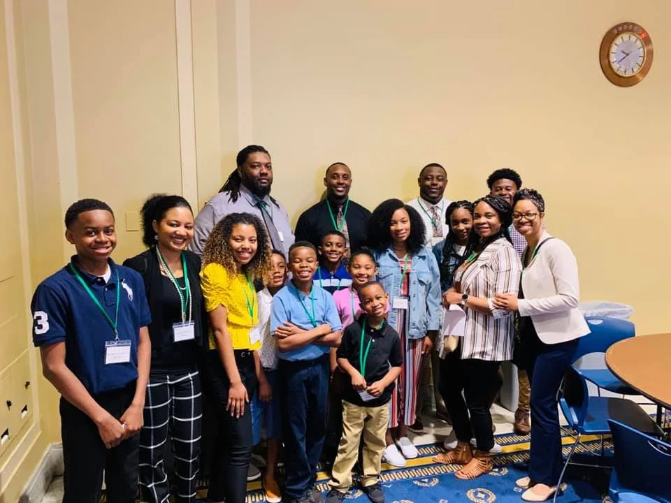 A diverse group of children and adults standing together in a conference room, smiling for the photo. They are wearing name tags and casual to business casual attire. There is a clock on the wall showing 10:10.