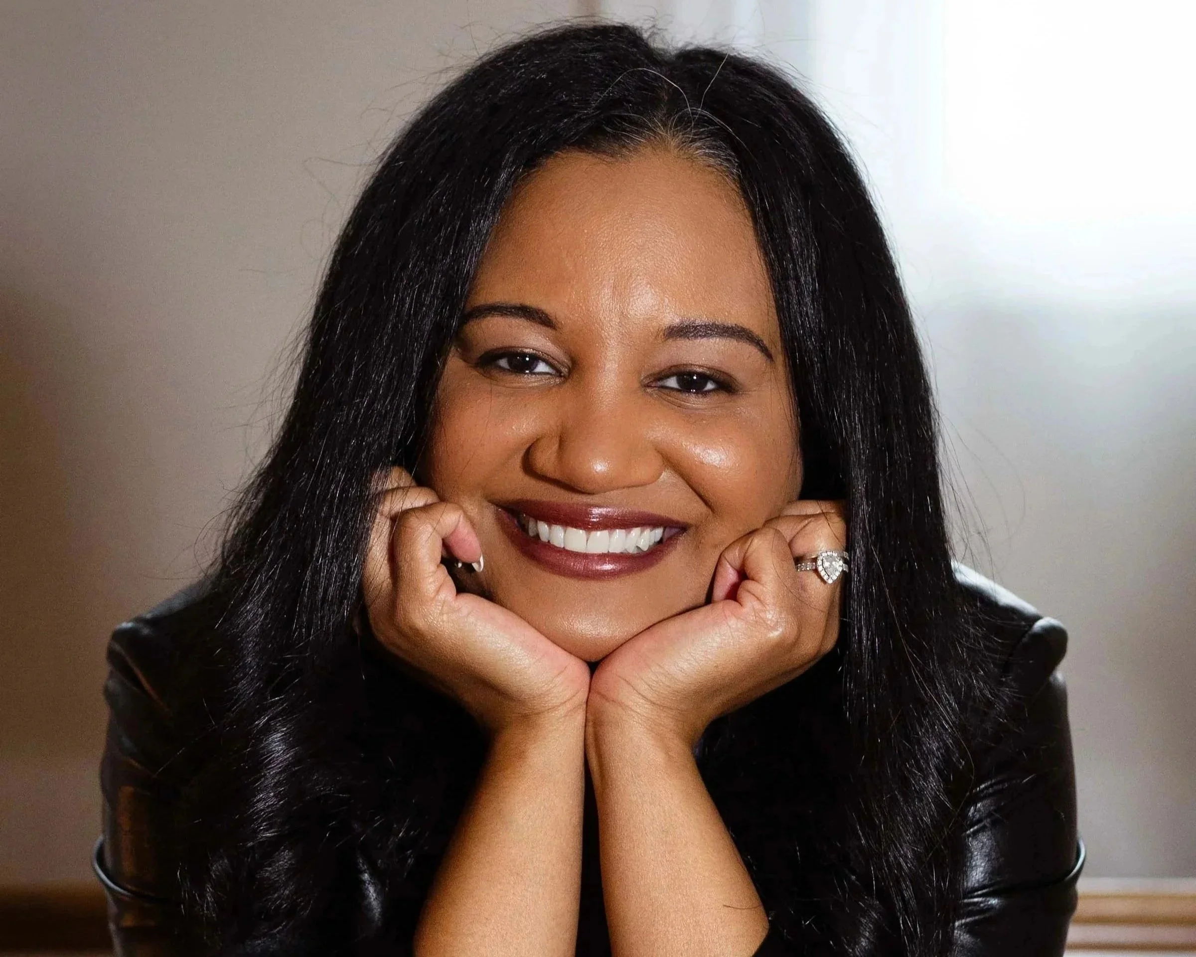 A woman with long black hair smiling and resting her chin on her hands, wearing a black leather jacket and a ring on her left hand.
