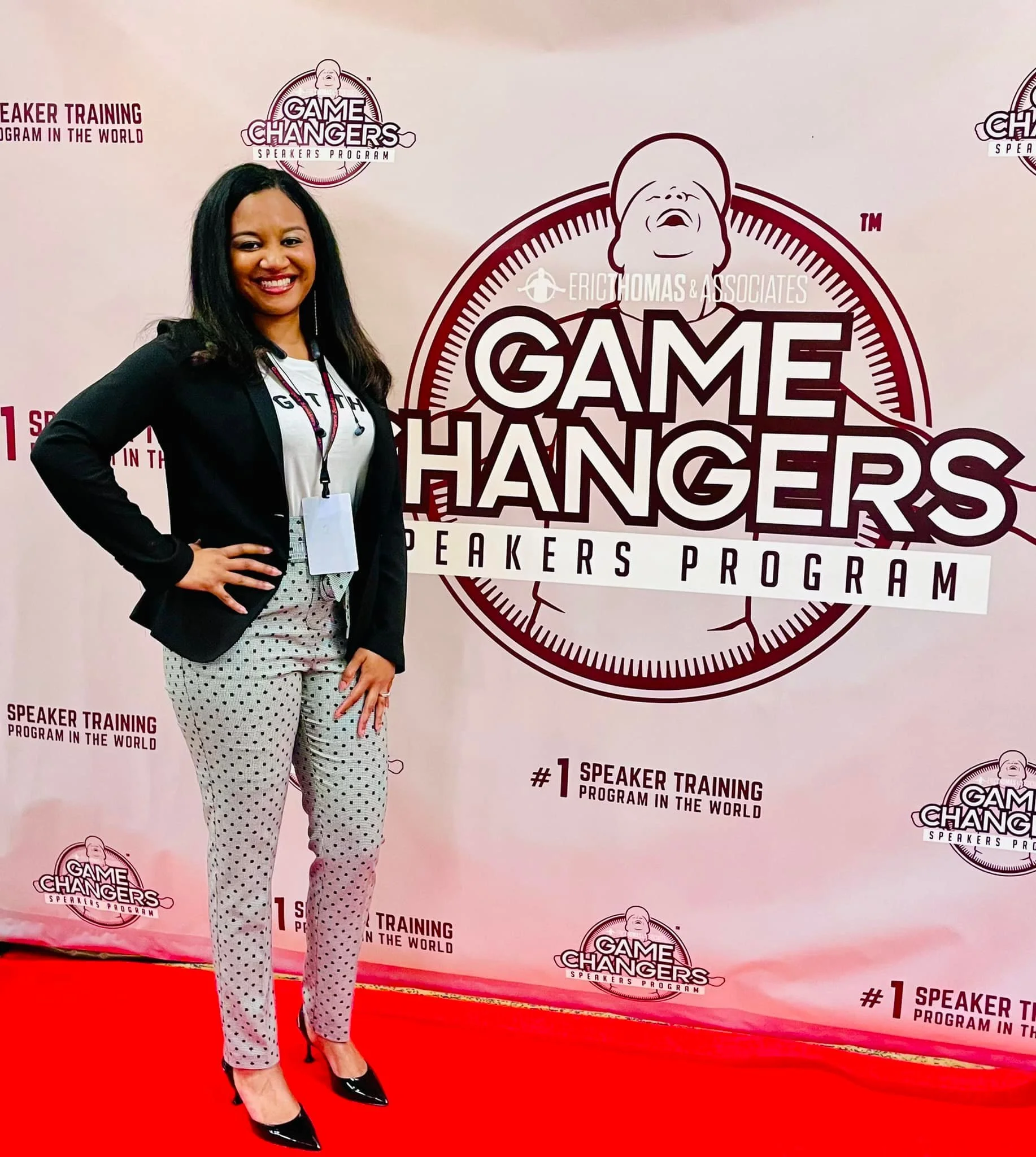 A woman standing on a red carpet at a conference, smiling, in business casual attire, with a lanyard and badge around her neck, in front of a backdrop featuring the 'Game Changers Speakers Program' logo.