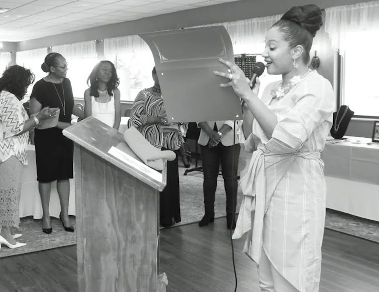 A woman lights a candle or takes a photo at a celebration or ceremony while others stand with their phones and watch. The woman is holding a microphone and standing near a podium in a well-lit room.