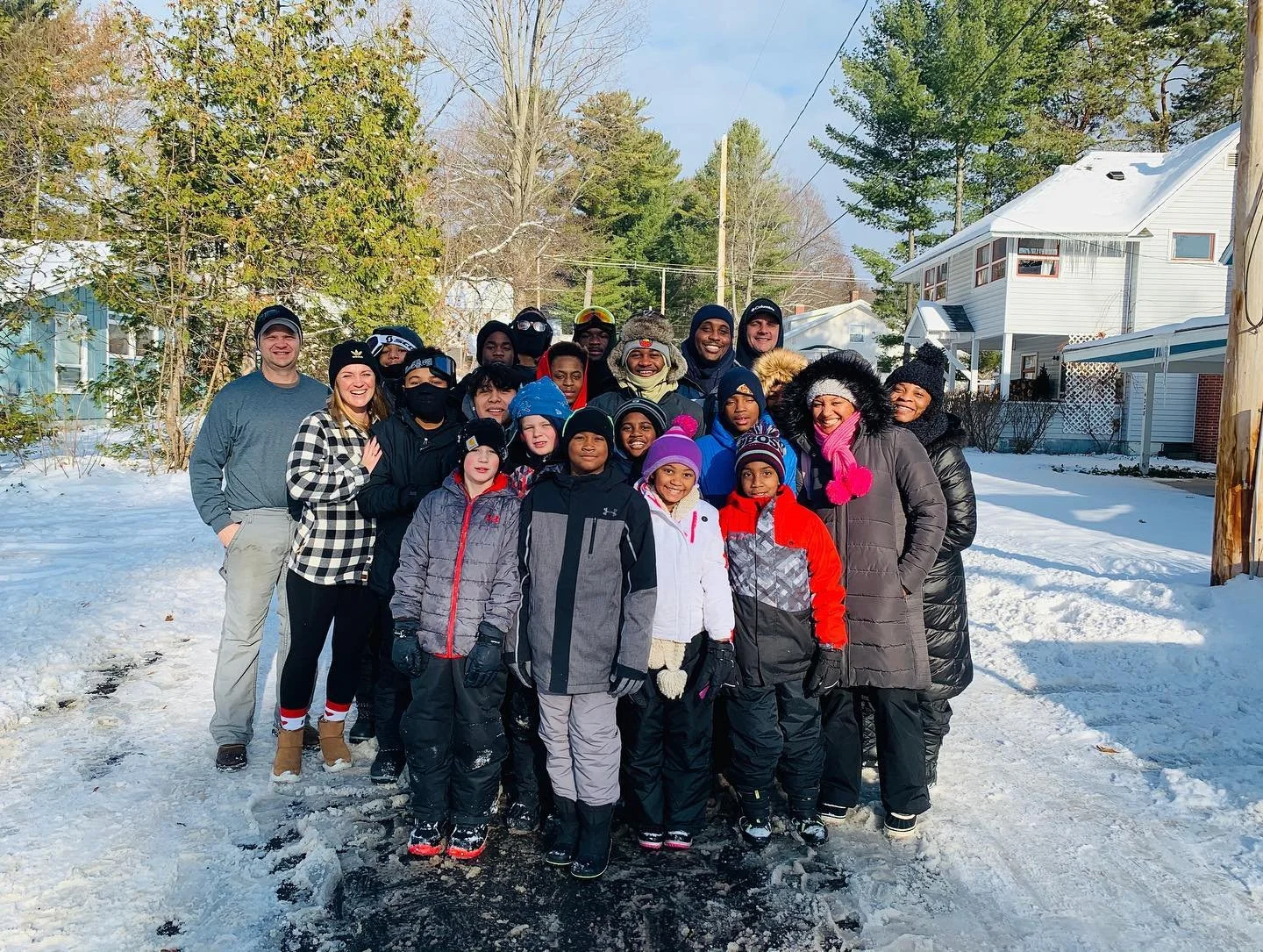 Group of children and adults standing outdoors in snowy weather, smiling for a photo in a residential neighborhood with trees and houses in the background.