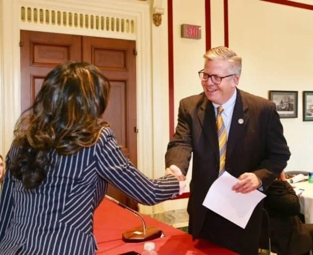 A man in a suit and glasses shaking hands with a woman with long dark hair and a striped blazer, in a room with wooden doors and framed pictures on the wall.