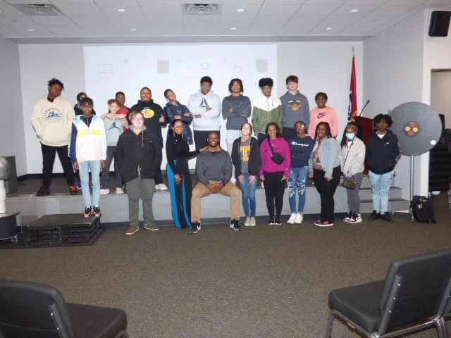 Group of diverse young adults posing on a stage in an indoor setting, with some people wearing masks and casual clothing, and a larger screen or projection behind them.