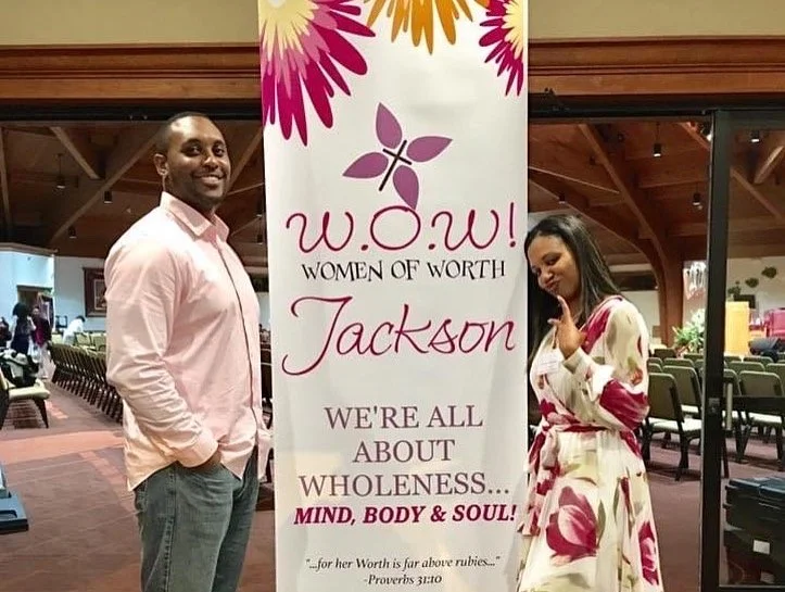 A man and woman standing next to a banner at an event. The banner reads, "w.o.w! Women of Worth Jackson. We're all about wholeness... mind, body, and soul!" The woman is wearing a floral dress and making a peace sign, while the man is wearing a light
