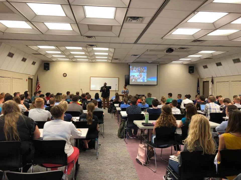 A classroom or conference room filled with students and a speaker at the front, presenting to an audience with a projected screen behind him.