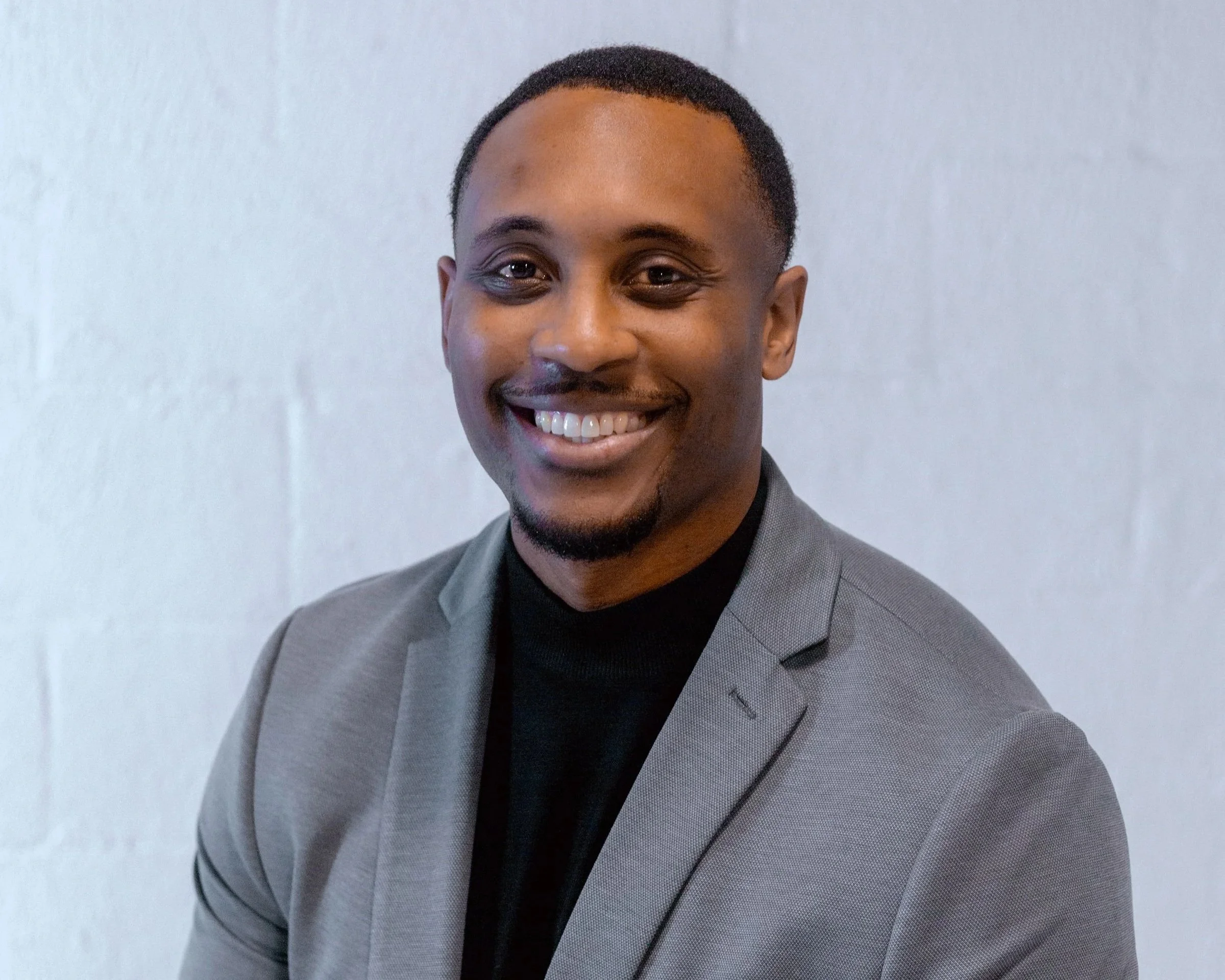 A smiling man with short dark hair, a light beard, and wearing a gray blazer over a black turtleneck, posing against a plain white wall.