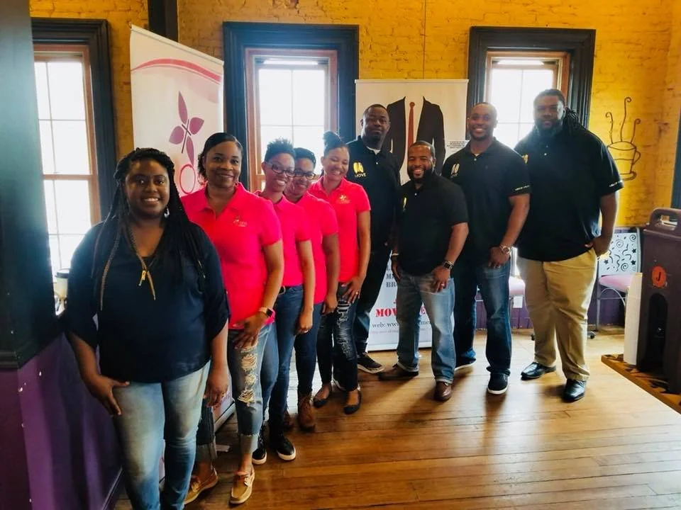 Group of nine people standing in a room with yellow brick walls, three windows, and wooden floors. Four women are wearing pink shirts, and five men are wearing black shirts. There are banners and artwork in the background.