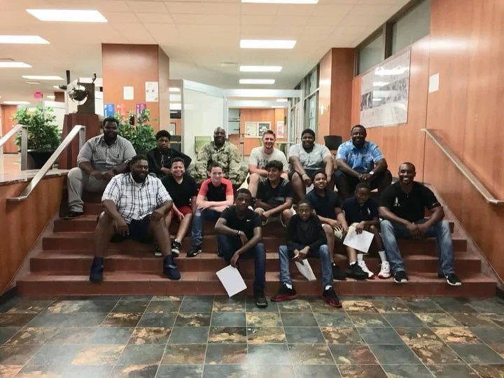 Group of 14 people, including adults and children, sitting on and around wooden stairs in an indoor setting, possibly a community center or building lobby.