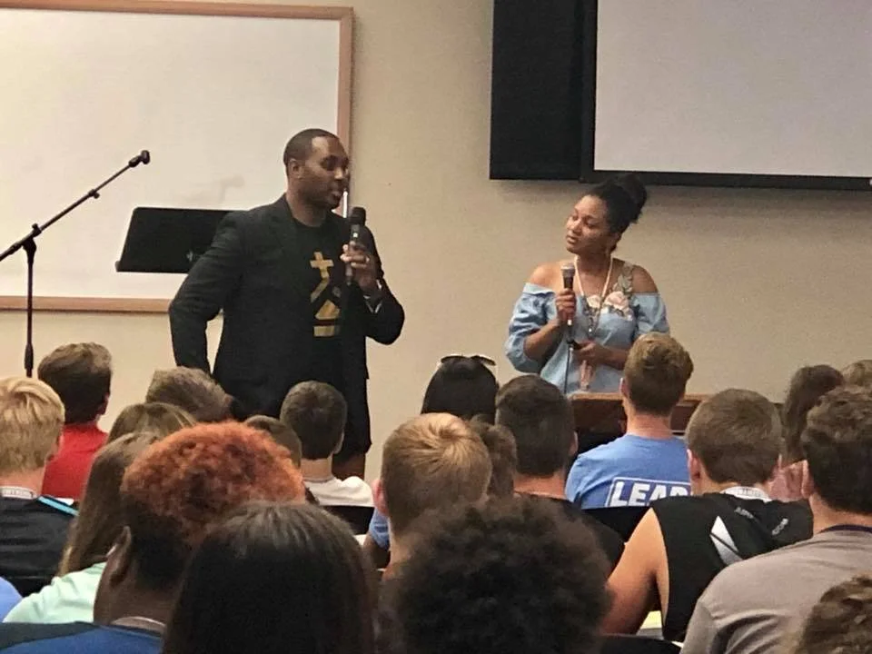 Two speakers, a man and a woman, are standing in front of an audience, holding microphones and speaking. The man is wearing a black blazer with a shirt, and the woman is wearing an off-shoulder blue dress. There is a podium and a whiteboard behind th