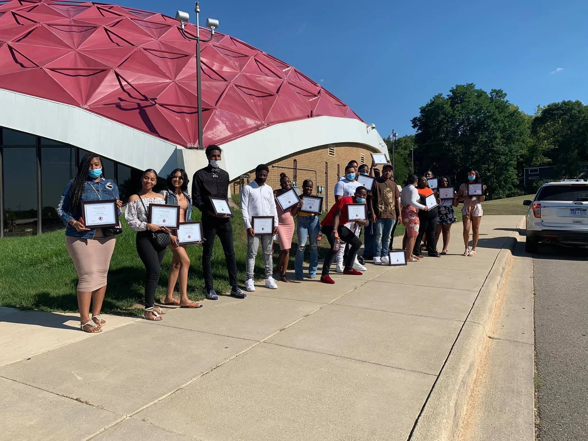 A group of young people standing outdoors on a sidewalk, holding framed certificates of achievement, with a distinctive pink and white building and green trees in the background. Some are wearing masks.