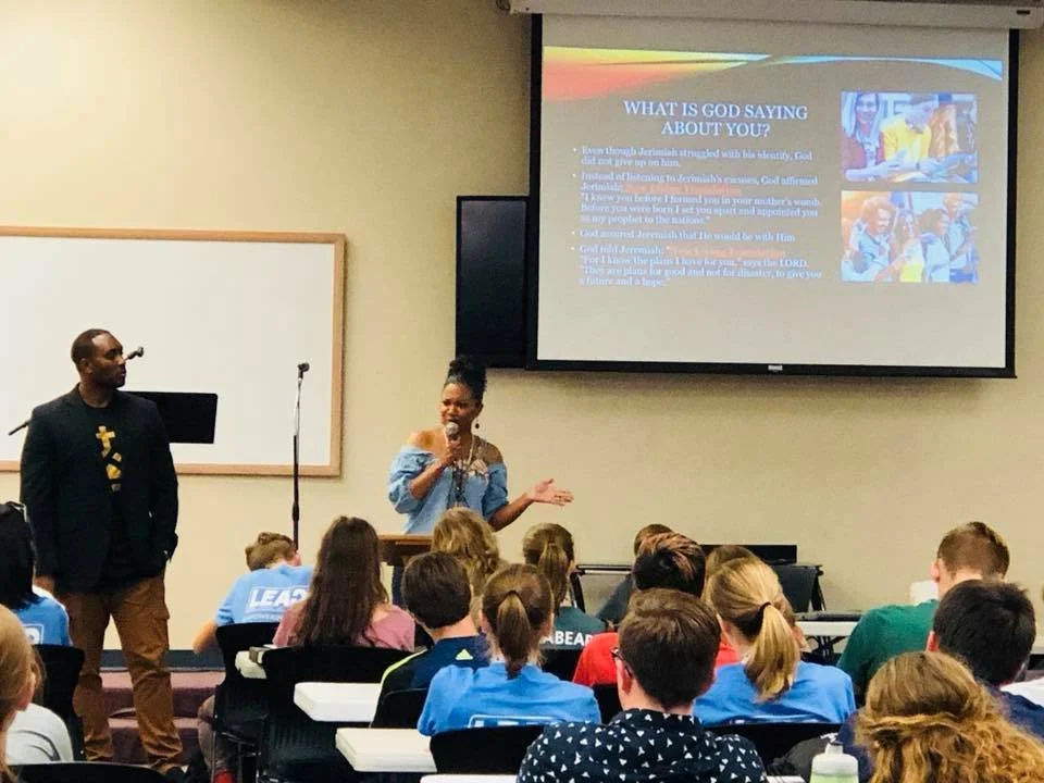 A classroom presentation with a woman speaking at the front, a man standing beside her, and students seated paying attention. The slide on the screen discusses what God says about you, with bullet points and images of children.