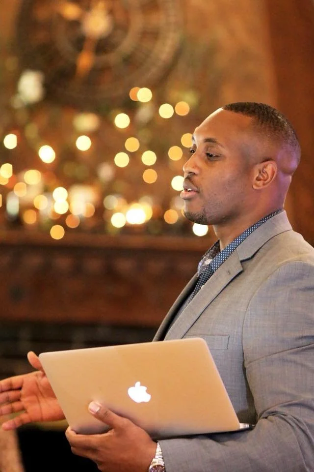 A man in a suit holding a closed Apple MacBook, looking to the side with a blurred background of warm lights.