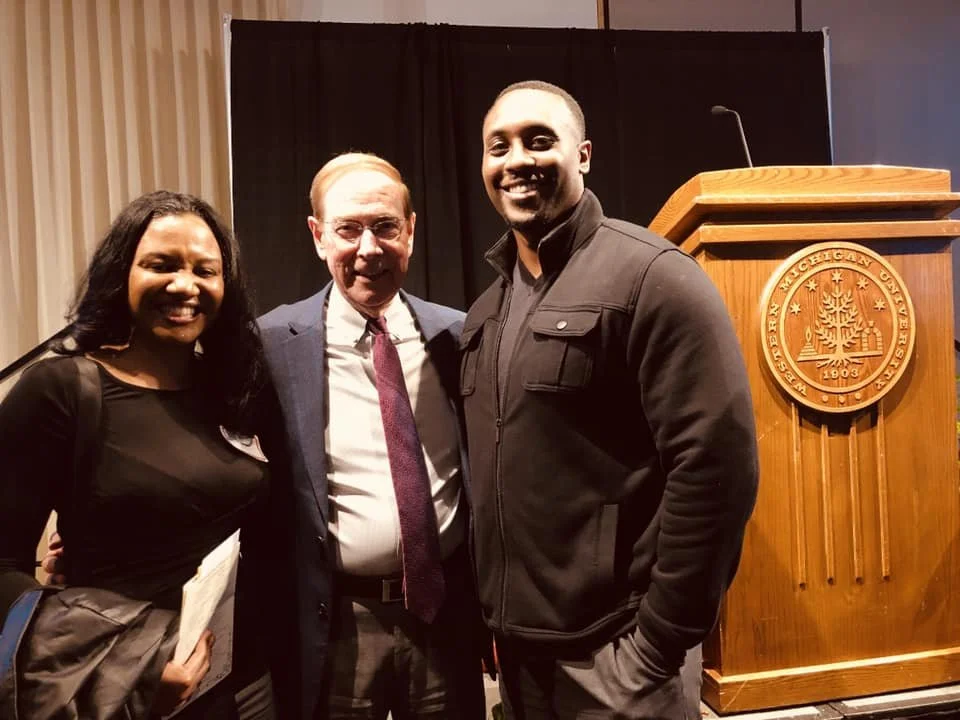 Three people standing together indoors near a wooden podium with a seal that reads 'Eastern Michigan University 1903'.