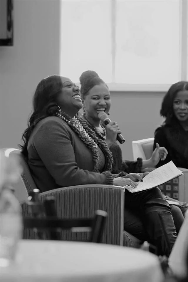 Women at a panel discussion or meeting, smiling and laughing, with one woman speaking into a microphone.