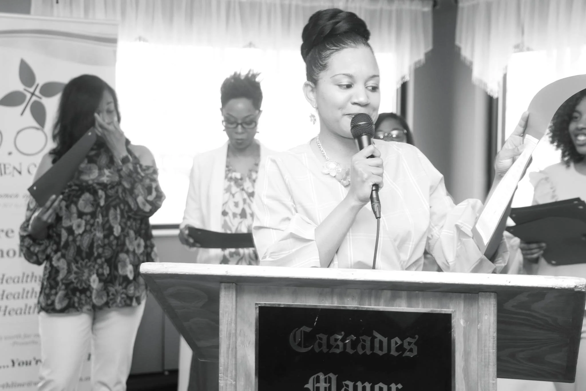 A woman speaking into a microphone at a podium with four women in the background during an event.