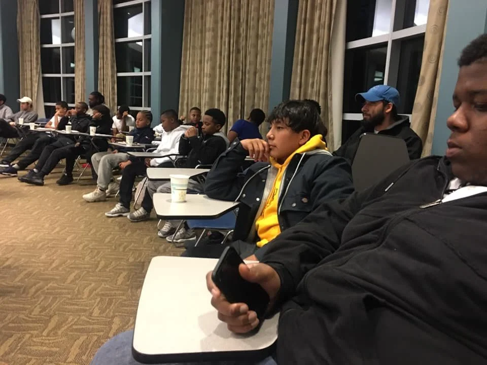 Group of children and teenagers sitting in a classroom or conference room, attentively listening, with some holding drinks and using electronic devices.