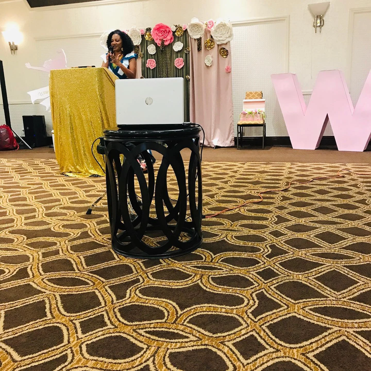 A woman speaking at a decorated event stage with large pink and white paper flowers and a pink backdrop. The stage has a golden cloth, and large pink letter 'W' on the right. There is a chair and a gift box on the stage.