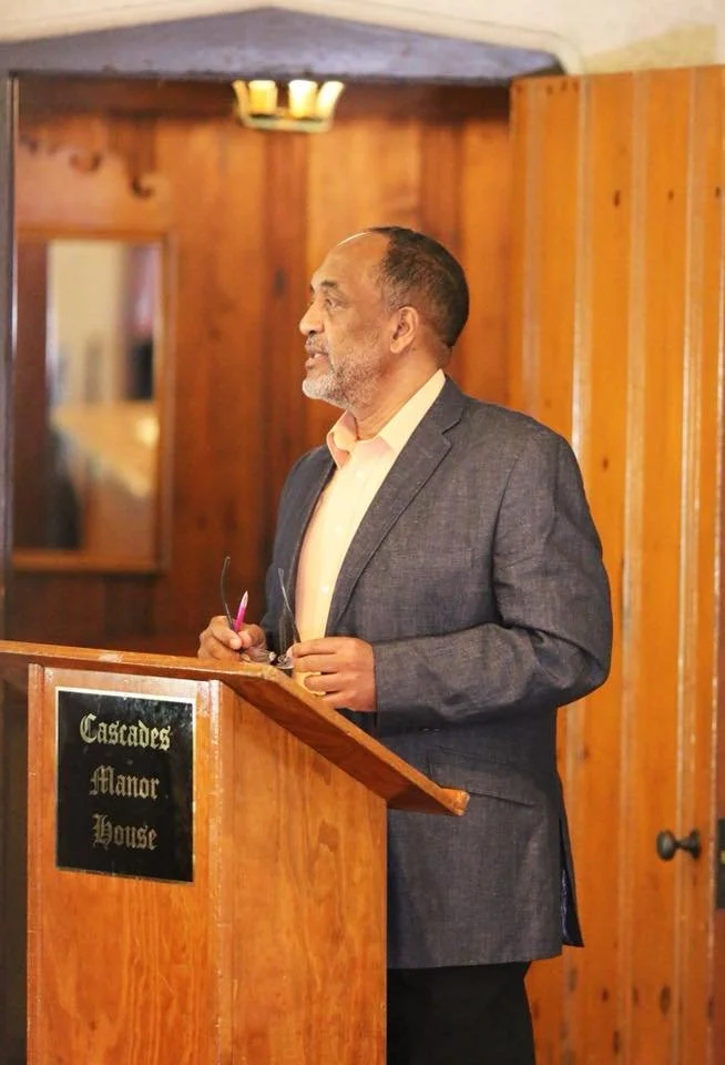 A middle-aged man with a beard, wearing a suit jacket and dress shirt, stands behind a wooden podium at Cascades Manor House, holding glasses and looking to the side in a warmly lit room.