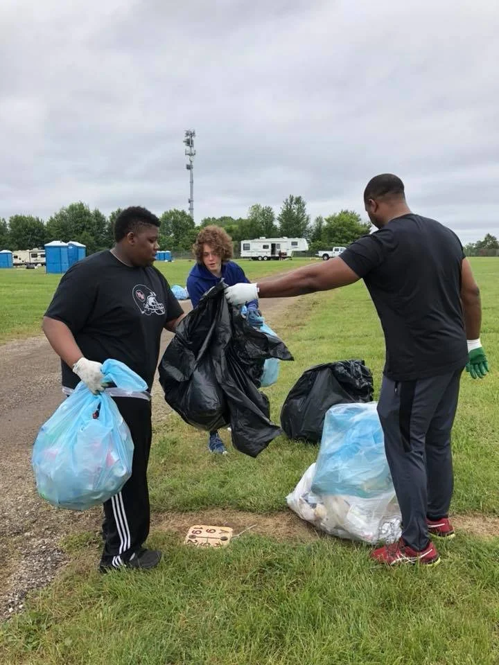 Three people are collecting trash outdoors, holding garbage bags and gloves, with RVs and portable toilets in the background.