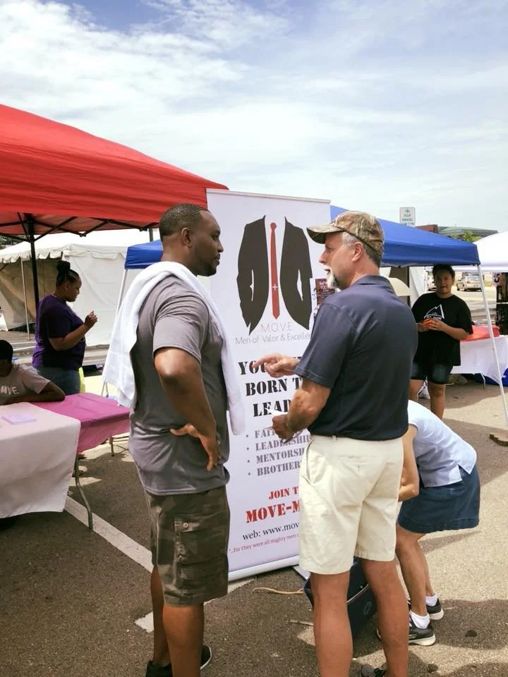 Two men are having a conversation at an outdoor event, standing in front of a banner with a suit and tie graphic. Several tents and other people are in the background.