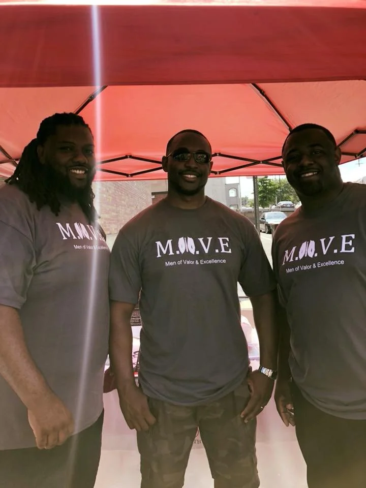 Three men standing under a red canopy, wearing matching gray T-shirts with the word "M.O.V.E" and the phrase "Men of Valor & Excellence," smiling at the camera.