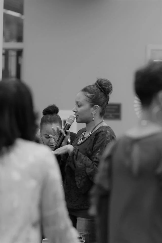 A woman in a black lace top is speaking into a microphone while holding a woman's hand, with a group of people around her. The scene appears to be indoors, possibly at a gathering or event.