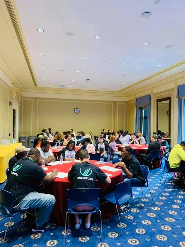 A large group of people seated at round tables in a conference room, engaged in discussions. The room has yellow walls, blue curtains, and a patterned blue carpet. Some people are wearing black shirts with a logo on the back.