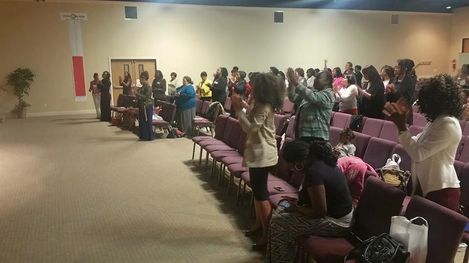 Group of women standing and clapping in a church or community room, some sitting in chairs, participating in a religious service or community event.