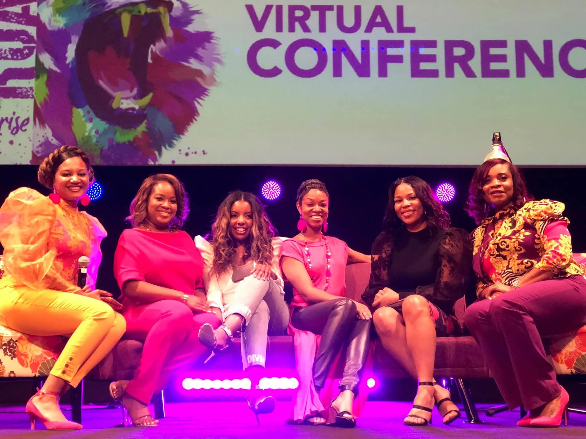 A group of seven women sitting on a couch on stage at a virtual conference, with a large screen behind them displaying a colorful lion and the words 'VIRTUAL CONFERENCE' in purple text.