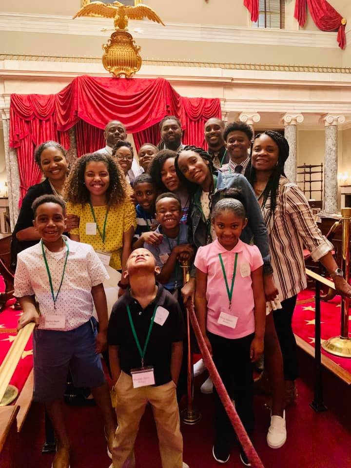 A group of children and adults posing together in a grand, ornate room with red drapes, marble columns, and gold accents.