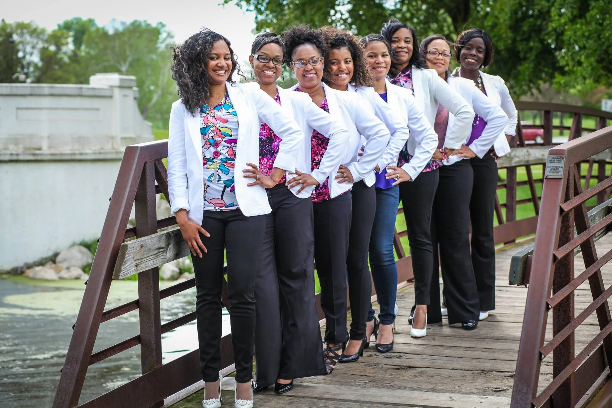 Seven women dressed in business casual attire, standing side by side on a wooden bridge outdoors, smiling at the camera.