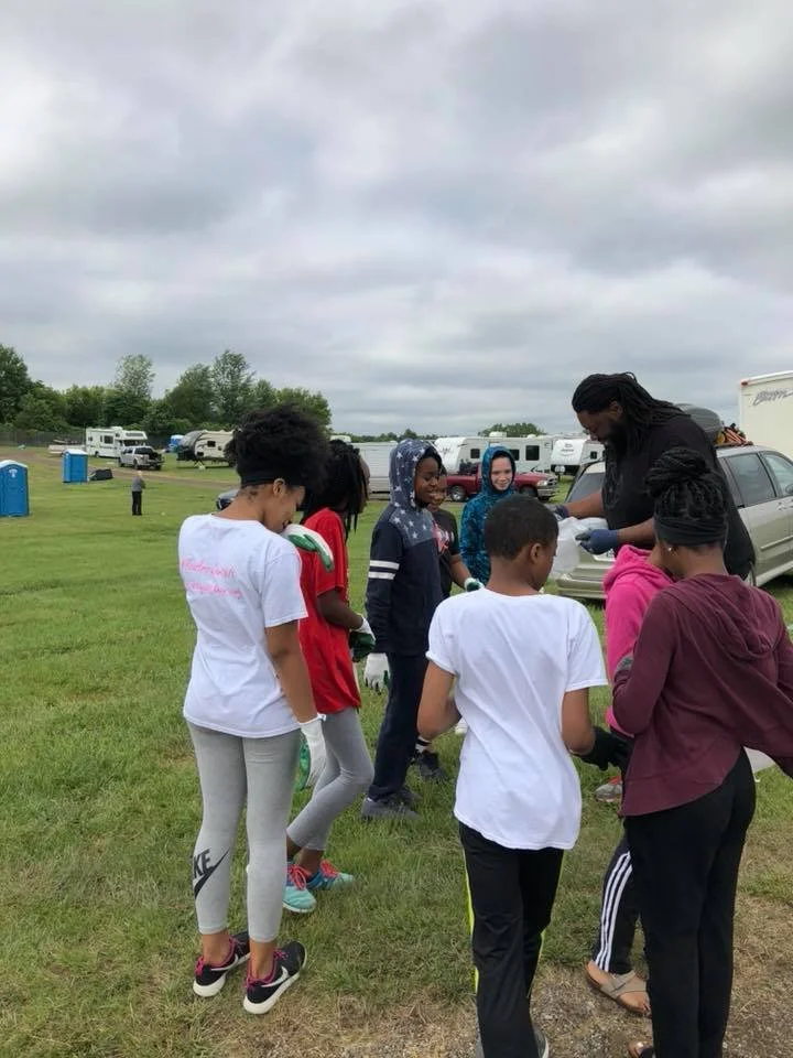 A group of children standing in line outdoors on a cloudy day, receiving food or supplies from an adult woman. The setting appears to be a park or open field with cars and campers in the background.