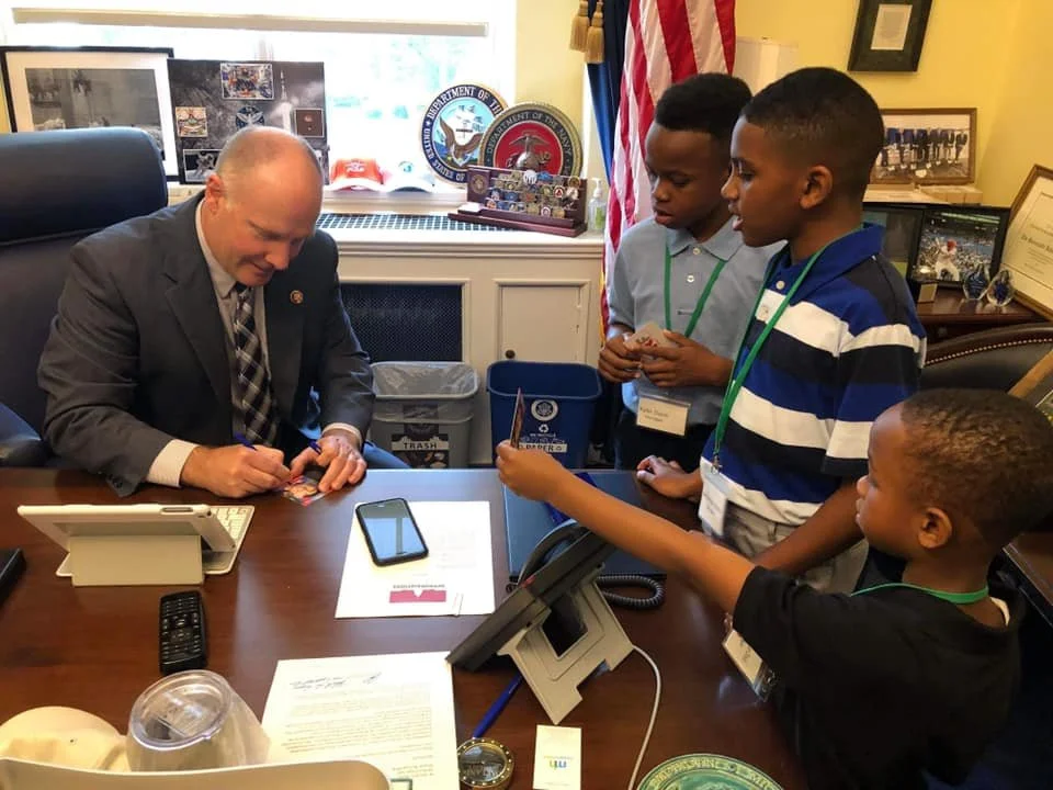 A man in a suit is sitting at a desk signing a photo for four boys who are standing around him, holding cards and looking at him. The setting appears to be an office with American flags, framed pictures, and seals related to the U.S. Department of Ve