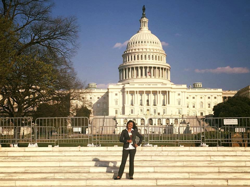 A woman in business attire standing on steps in front of the US Capitol building in Washington, D.C. with cloudy blue sky background.