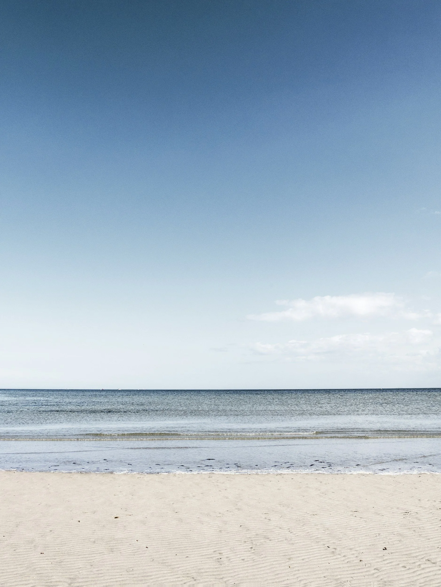 A sandy beach with shells and pebbles, calm ocean water, and a clear sky with few clouds.
