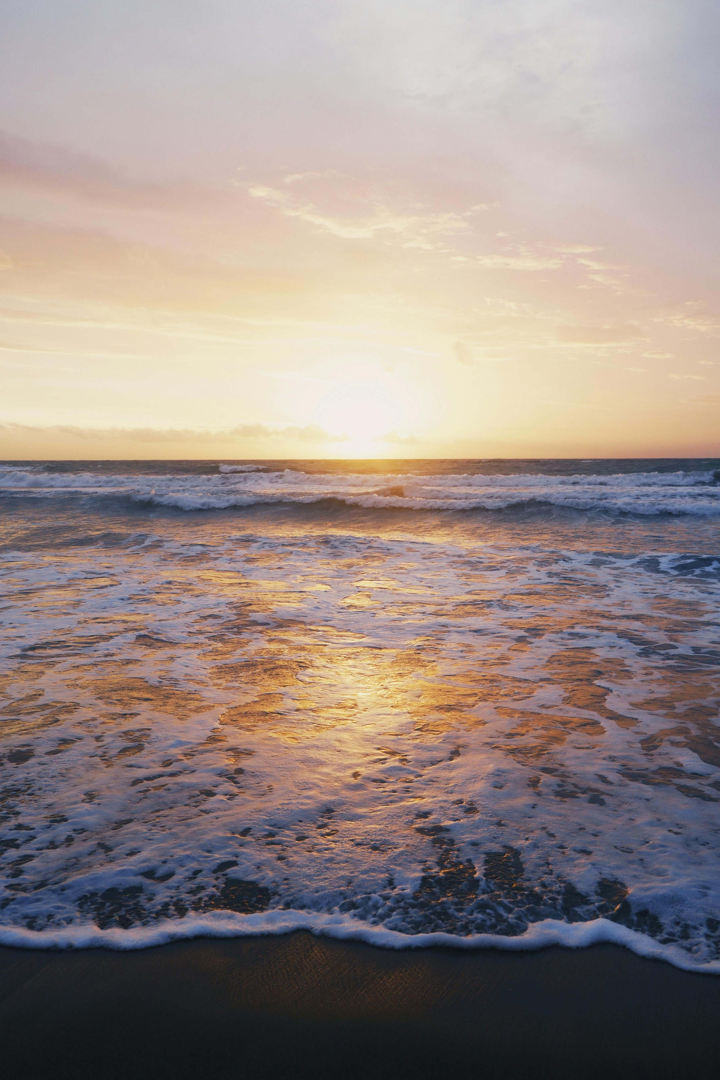 Sunset over the ocean with waves crashing onto the shore and a partly cloudy sky.